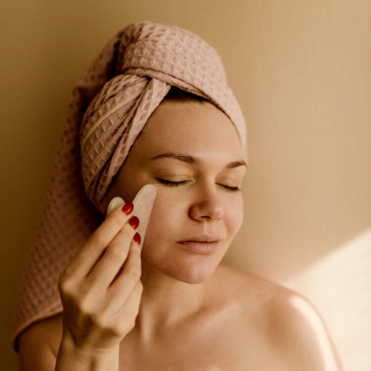 Mulher com uma toalha na cabeça utilizando a pedra Gua Sha para massagear o rosto.